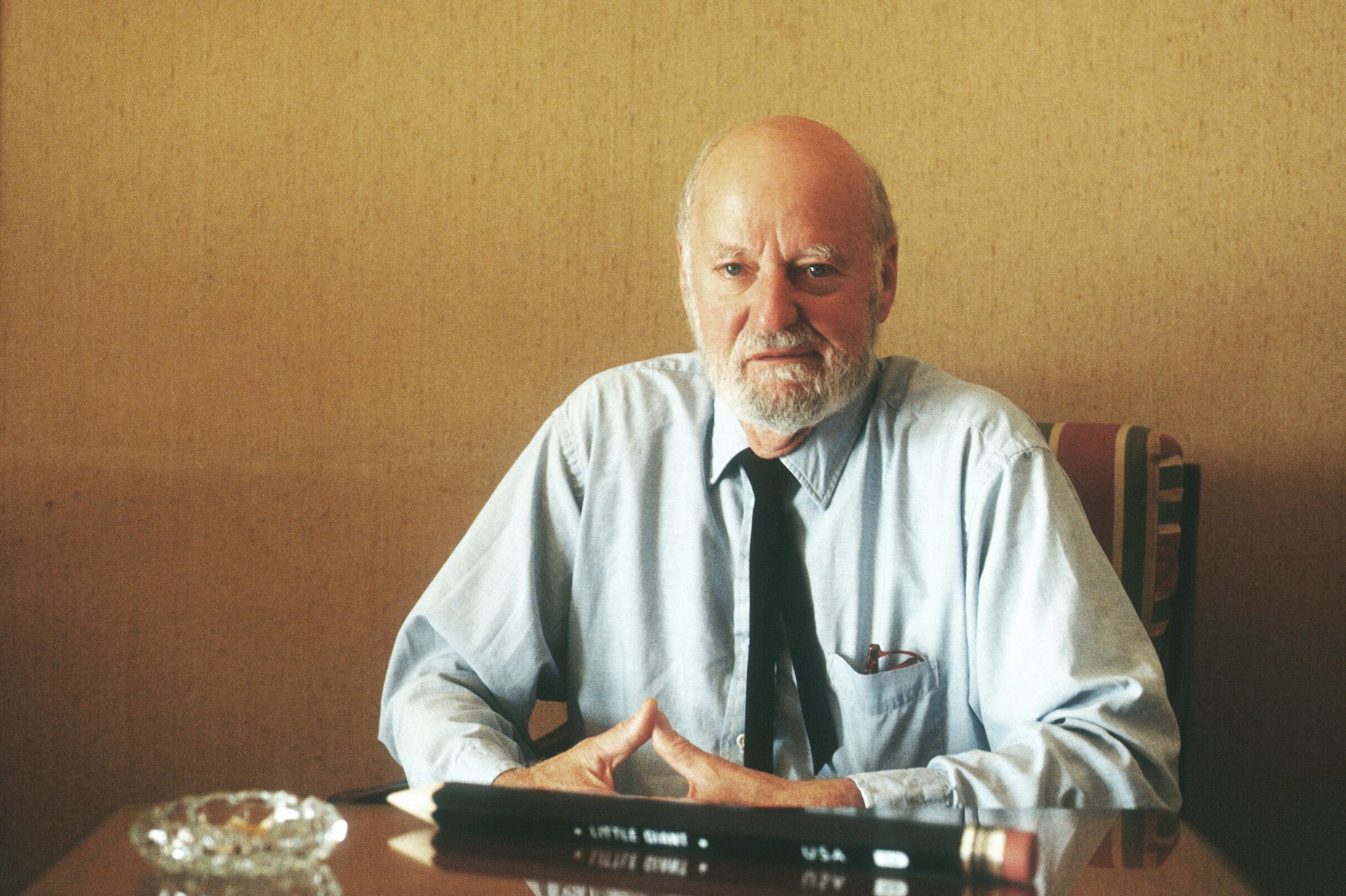 Lawrence Ferlinghetti sitting with a giant pencil and an ash tray in front of him